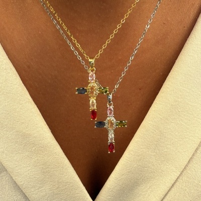 Dois colares com pendentes cruz gemas coloridas, correntes dourada e prateada em pele vestindo roupa branca.
