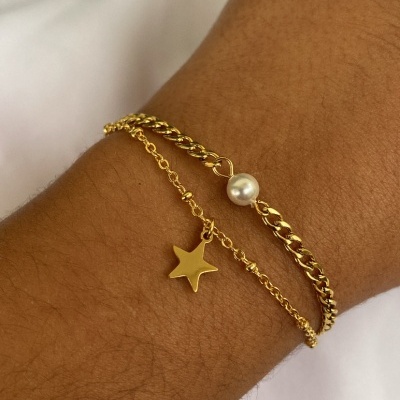 Pulseira dourada dupla com estrela e pérola num pulso sobre tecido branco
