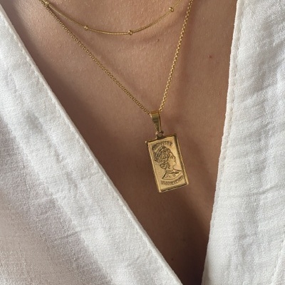 Colar duplo dourado com pendente retangular gravado, sobre blusa branca de linho.