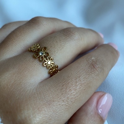 Anel dourado com flores e pedra clara no dedo médio, unhas rosa claro, fundo branco