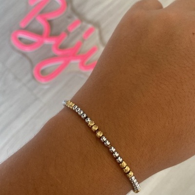 Pulseira fina dourada e prateada num pulso com fundo claro e texto rosa desfocado