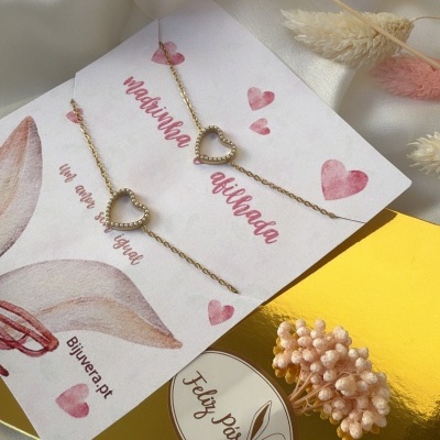 Pulseiras douradas com coração e embalagem com textos em cartão branco sobre tecido acetinado creme e flores secas rosa e creme