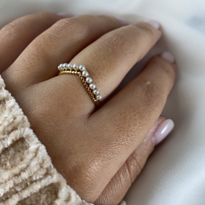 Anel dourado com pérolas pequenas no dedo médio, mão com unhas pintadas, fundo de tecido branco e malha bege.