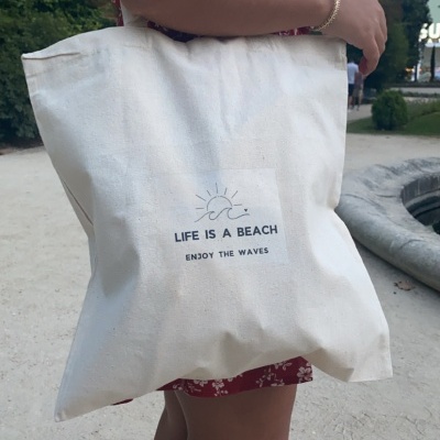 Bolsa de tecido creme com texto 'LIFE IS A BEACH ENJOY THE WAVES' e desenho de sol e ondas, usada por pessoa com vestido vermelho de flores brancas.