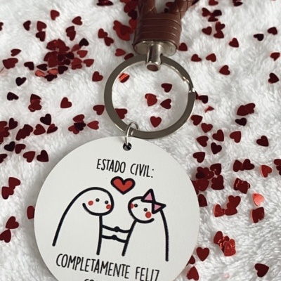 Chaveiro redondo branco com desenho de casal e texto em português sobre superfície branca com confetes vermelhos.