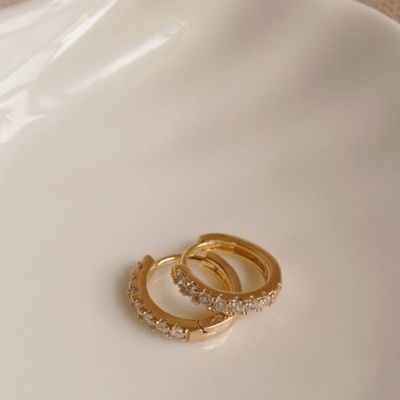 Brincos de argola dourados com pedras brancas numa superfície branca lisa