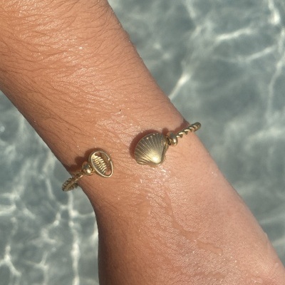 Pulseira dourada com pingentes de concha num pulso sobre água cristalina