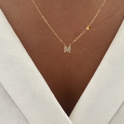 Colar dourado com letra M em cristais e coração, usado no pescoço sobre vestido branco