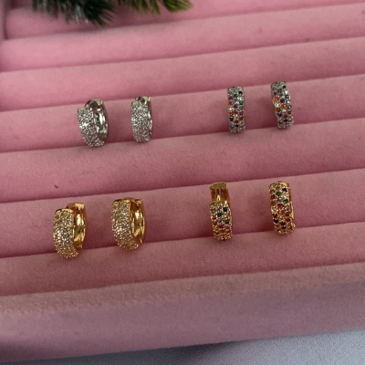 Oito pares de brincos em metal prateado e dourado com pedras brilhantes, organizados em suporte de veludo rosa.