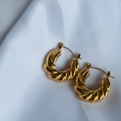 Brincos de argola dourados com padrão ondulado sobre tecido branco.