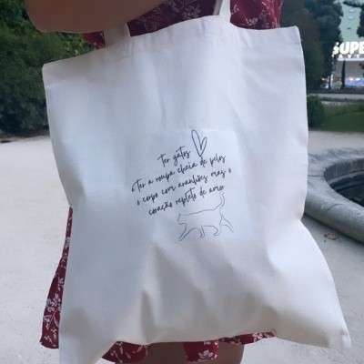 Mala de tecido branco com ilustração e texto sobre gatos, segurada por mulher com vestido vermelho floral, em ambiente exterior