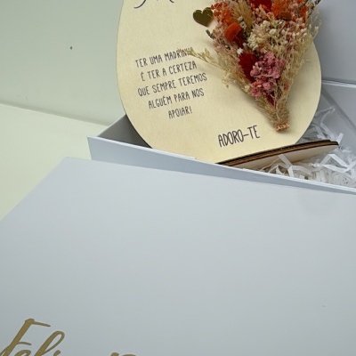 Embalagem branca aberta com decoração de madeira em forma de ovo com flores secas e texto dedicado a madrinha.