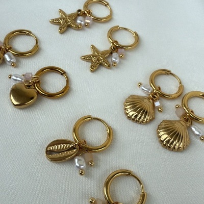 Brincos dourados com pingentes de concha, estrela-do-mar, coração e búzio com pérolas sobre tecido branco