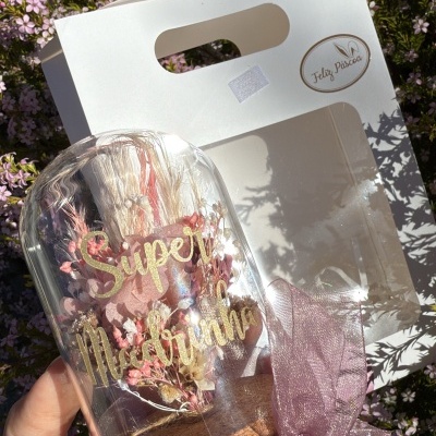 Vaso decorativo de vidro com flores secas e texto 'Super Mãe' em frente a saco branco com texto 'Feliz Páscoa'