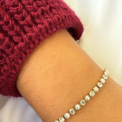 Pulseira tennis com pedras brancas montada em metal dourado no pulso com manga vermelha de malha.