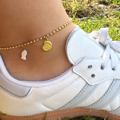 Ténis brancos com pulseira dourada no tornozelo, relvado e flores no fundo.