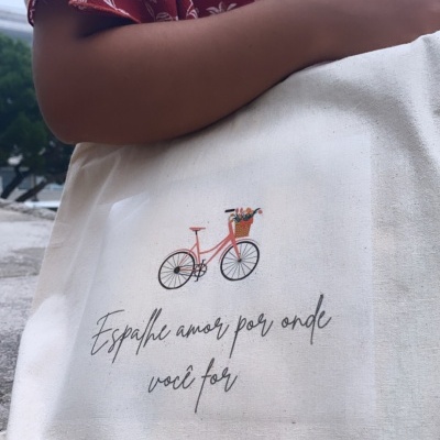 Saco de tecido com estampa de bicicleta e texto, segurado por pessoa de vestido vermelho de flores