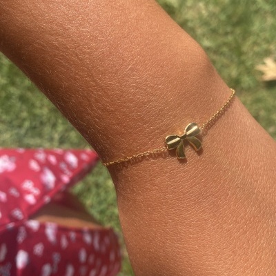 Pulseira dourada com pendente em forma de laço no pulso, fundo com relva e roupa vermelha com estampas brancas