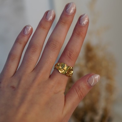 Anel dourado com textura ondulada no dedo anelar de uma mão com unhas pintadas de rosa claro com linhas brancas