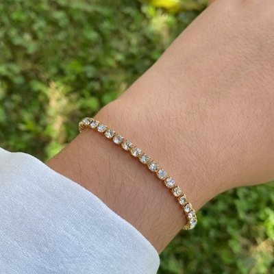 Pulseira tennis dourada com pedras transparentes no pulso em fundo de relva