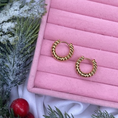 Brincos dourados em argola torcidos sobre almofada rosa com decoração de ramos verdes e bagas vermelhas.