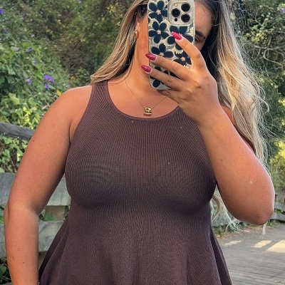 Top feminino de malha castanha com gola redonda e corte evasê, usado com calças de ganga azuis, selfie ao ar livre.