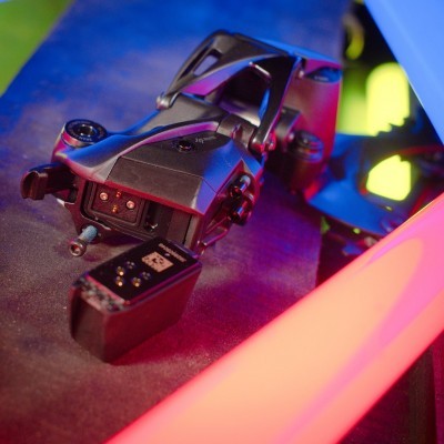 Drone preto com bateria removível em superfície escura iluminada em azul e rosa