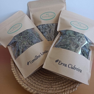 Três embalagens de papel kraft com ervas secas visíveis, etiquetadas Tomilho Limão e Erva Cidreira, sobre suporte de palha.