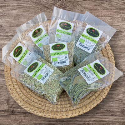 Seis pacotes com ervas secas Bio Aroma Natural numa tábua e tabuleiro de palha sobre mesa de madeira.