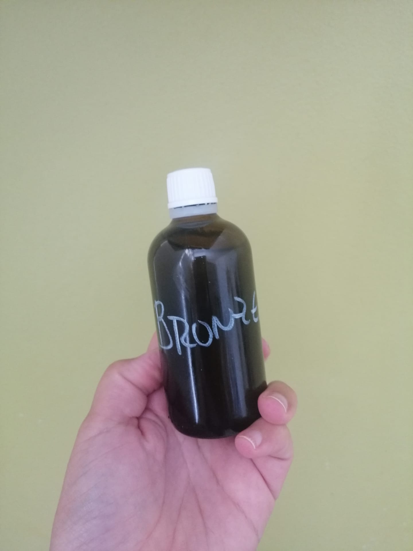 Frasco castanho com tampa branca e texto 'BRONZE' segurado numa mão, fundo verde-claro