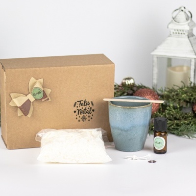 Conjunto de presente com caixa de cartão decorada, vela azul, frasco de óleo essencial e saco branco, com decoração natalícia ao fundo.