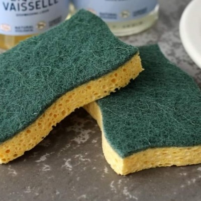 Duas esponjas de lavar amarelas com superfície verde abrasiva sobre bancada cinza com garrafas ao fundo