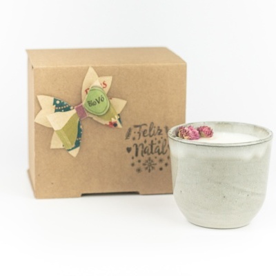 Vela branca em cerâmica cinzenta com flores secas e caixa de cartão com laço e texto 'Feliz Natal'