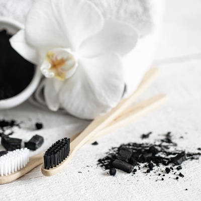 Duas escovas de dentes de madeira com cerdas preta e branca, carvão ativado em pó e pedaços, e flor branca sobre fundo claro