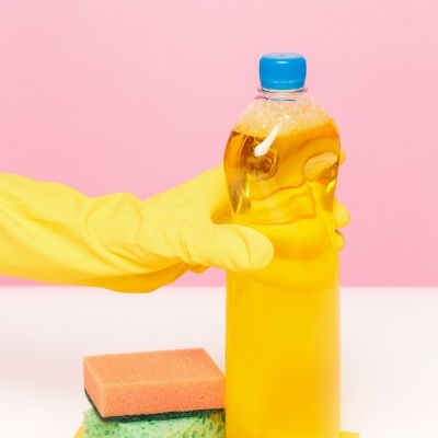 Garrafa plástica com líquido amarelo segurada com luva amarela, esponjas e pano, fundo rosa.