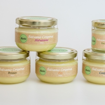 Cinco frascos de bálsamo corporal Balvô em vidro com tampas douradas e etiquetas verdes e creme sobre fundo claro.