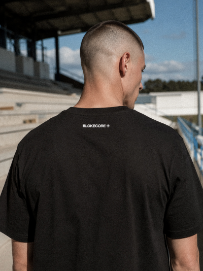 T-shirt preta com texto branco BLOKECORE + nas costas, vista de trás em ambiente exterior
