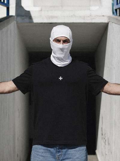 Homem com balaclava branca e t-shirt preta com símbolo branco no peito num túnel de cimento