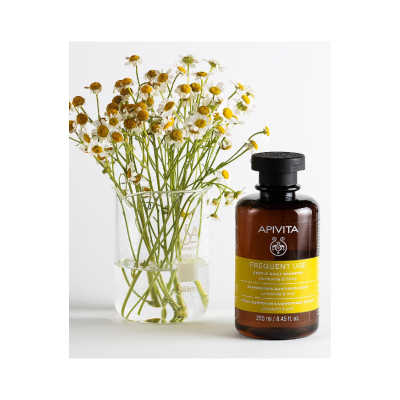 Frasco de champô APIVITA com flores de camomila em copo de vidro.