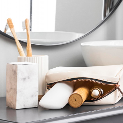 Acessórios de higiene numa casa de banho com escovas de dentes de madeira numa jarra branca, caixa marmoreada, saboneteira branca e necessaire creme aberta com itens dentro.