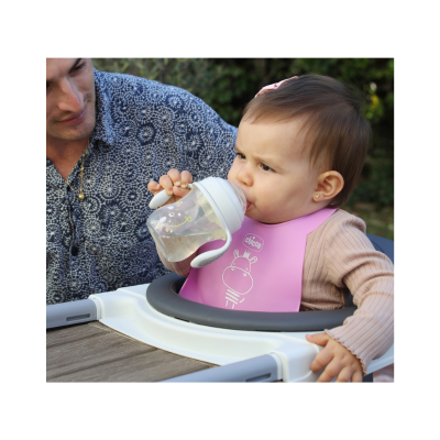 Bebé a beber de copo com bico, usa babete rosa Chicco, sentado em cadeira alta junto a mesa de madeira