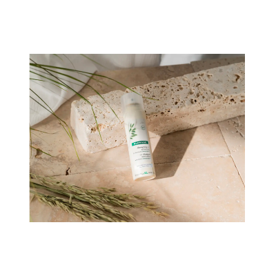 Frasco de shampoo a seco Klorane sobre superfície de pedra bege com ramos verdes