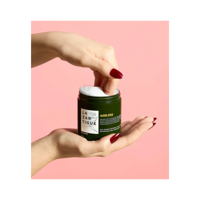 Frasco de creme LAZARTIGUE AGELESS sendo segurado por mãos femininas com unhas vermelhas sobre fundo rosa.