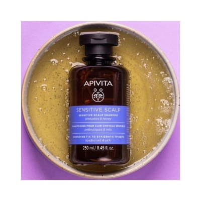 Frasco castanho de champô APIVITA Sensitive Scalp com etiqueta azul numa base redonda com líquido amarelo