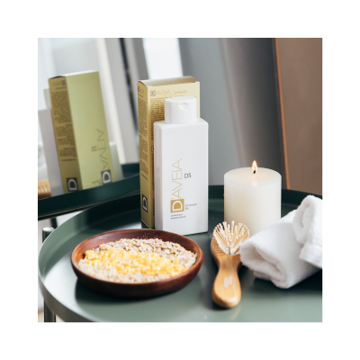 Frasco branco de champô DAVELA DS com embalagem dourada, tigela com flocos de aveia, escova de madeira, toalha branca e vela acesa numa mesa verde.