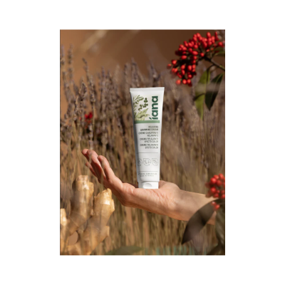 Tubo branco de creme relaxante Iana segurado por uma mão em fundo natural com plantas e frutos vermelhos