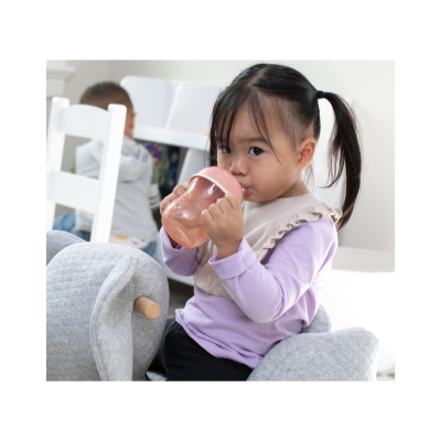 Criança sentada a beber de um copo rosa numa cadeira de tecido cinza acolchoada.