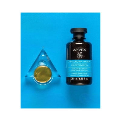 Frasco preto de shampoo APIVITA HYALURONIC HYDRA com etiqueta azul claro e objeto triangular translúcido com líquido dourado sobre fundo azul