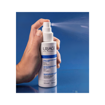 Frasco branco de spray Uriage Bariéderm-CICA a ser pulverizado na mão contra fundo azul.