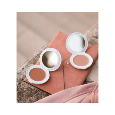 Dois pós compactos em embalagem branca com espelhos e uma carteira castanha clara sobre pedra.